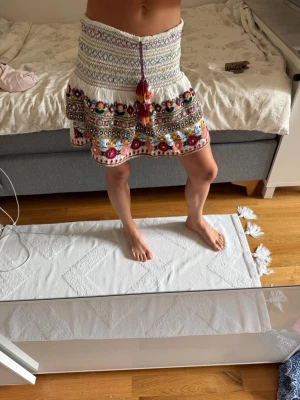 Vit kortkjol med broderade detaljer - Supersöt vit kortkjol med färgglada broderier och volangkant. Kjolen har ett boho-inspirerat mönster, snörning med tofsar framtill och sitter högt i midjan. 