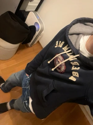 Mörkblå hoodie från Abercrombie - Mörkblå hoodie från Abercrombie med stor huva och grått innerfoder. Broderad logga och text på bröstet, vita dragsnören och känguruficka framtill. Perfekt för en chill och avslappnad stil.