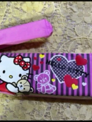 Hello Kitty plånbok med nallemotiv - Söt plånbok med Hello Kitty och nallemotiv på framsidan. Plånboken har lila och rosa ränder, dekorerad med hjärtan och texten 'Hello Kitty'. Praktisk dragkedja och handrem i rosa tyg. Perfekt för dig som gillar gulliga accessoarer.