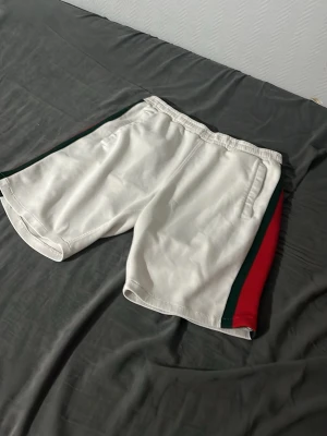 Vita shorts med röd och grön rand - Säljer ett par vita shorts med breda ränder i rött och grönt längs sidorna. Shortsen har elastisk midja och sidofickor, perfekta för en avslappnad och sportig stil. Materialet känns mjukt och bekvämt, troligen bomull eller polyester.