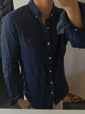 Mörkblå linneskjorta från Polo Ralph Lauren - Klassisk mörkblå skjorta i linne från Polo Ralph Lauren med vit knappstängning och röd broderad logga på bröstet. Skjortan har lång ärm och button-down krage, perfekt för en clean och stilren look. 100% linne