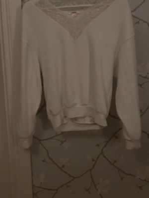 Vit sweatshirt med spetsdetalj  - Säljer en vit sweatshirt med bred mudd i midjan och vid ärmsluten. Tröjan har en snygg V-ringning med spetsdetalj upptill som ger en unik look. Perfekt för dig som gillar stilrena plagg med en twist.