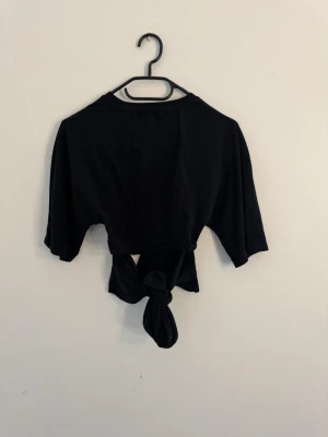 Svart croppad topp med knyt - Säljer en svart croppad topp med justerbar rosett bak på ryggen längst ner. Toppen har korta ärmar och rund halsringning. Perfekt för dig som gillar en enkel men trendig stil. Materialet känns mjukt och bekvämt. Aldrig använd.