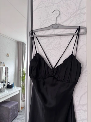 Svart långklänning med smala band - Elegant svart långklänning i satinliknande material med smala axelband som korsas i ryggen. Den är perfekt till bal eller fina tillställningar med en slits, 48 cm lång. Skriv om du undrar över mått💕
