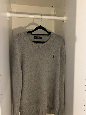 Grå stickad tröja från Polo Ralph Lauren - Klassisk grå stickad tröja från Polo Ralph Lauren med rund halsringning och diskret broderad logga på bröstet. Tröjan har långa ärmar och ribbade muddar vid ärmslut och nederkant. Perfekt basplagg i garderoben.