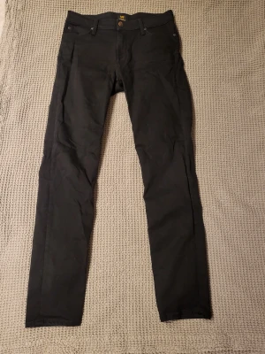 Svarta slim fit jeans från Lee - Snygga svarta slim fit jeans från Lee med klassisk femficksdesign.. Jeansen har raka ben och är tillverkade i ett mjukt, stretchigt material som ger en bekväm passform. Perfekta för dig som gillar en stilren och enkel look.