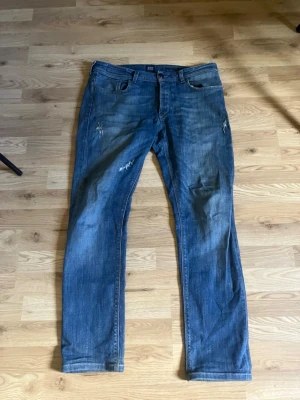 Boggi milano jeans - Säljer ett par blå jeans med raka ben och slitningar på låren och knäna. Jeansen har klassisk femficksdesign och är gjorda i denim. Perfekta för en avslappnad och trendig look.