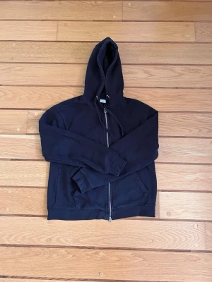 Svart hoodie med dragkedja från Zara - Säljer en svart hoodie från Zara med dragkedja framtill och två fickor. Tröjan har huva med dragsko och ribbade muddar vid ärmslut och nederkant. 