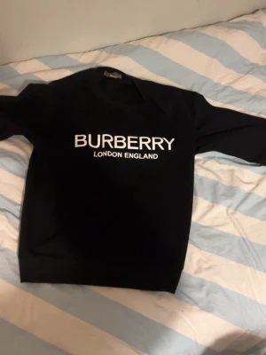 Svart Burberry sweatshirt - Svart sweatshirt från Burberry med vit logga och texten 'London England' på bröstet. Klassisk rund halsringning och långa ärmar. Perfekt för en stilren och trendig look.
