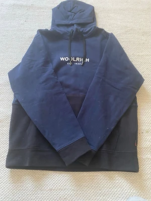 Marinblå och svart hoodie från Woolrich - Snygg marinblå hoodie från Woolrich med svart nederdel och ficka framtill. Tryckt logga på bröstet och klassisk huva med dragsko. Perfekt för en avslappnad streetwear-look. Mjuk bomullskänsla och ribbade muddar vid ärmslut.