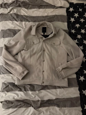 Ljusgrå höstjacka från New Look - Snygg ljusgrå höstjacka från New Look med klassisk krage, bröstfickor med lock och knappar framtill. Jackan har raka sömmar och en stilren look som passar perfekt till många outfits. Enkel att matcha och riktigt trendig.