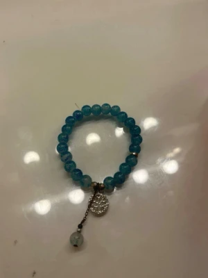 Blått pärlarmband med berlock - Snyggt armband med blå pärlor och elastiskt band. Armbandet har en silverfärgad berlock med livets träd och en extra hängande pärla. Perfekt för dig som gillar detaljer och vill addera lite färg till din stil. Missfärgad, se bättre på bild 2