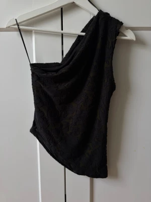 Svart oneshoulder topp med struktur - Snygg svart topp med oneshoulder-design och smal axelrem. Tyget har en diskret strukturerad yta med blommönster som ger en cool touch. Perfekt för dig som vill sticka ut lite extra med en unik topp.