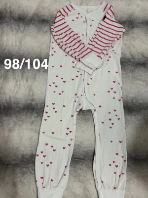 Vit pyjamas med rosa hjärtan - Mysig vit pyjamas med rosa hjärtan och långärmade ärmar med rosa ränder. Helknäppt framtill och muddar vid benslut för extra komfort. Perfekt för sköna hemmakvällar och sovstunder.🌸 Fint använt skick. Storlek 98/104