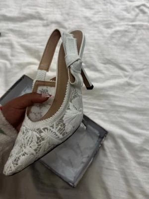 Vita spetsiga pumps med rosett - Säljer ett par eleganta vita pumps med spetsdetaljer och öppen häl. Skorna har en fin rosett på remmen och spetsig tå. Klacken är smal och ger en klassisk look. Perfekta för dig som vill ha något extra till outfiten.