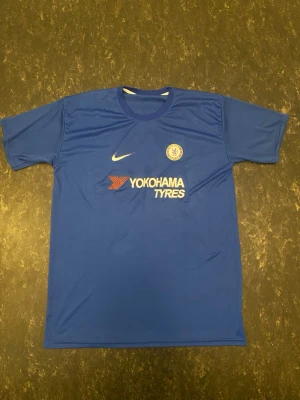 Chelsea matchtröja Hazard 10 Nike - Blå Chelsea fotbollströja med tryck 'Hazard 10' på ryggen och 'Yokohama Tyres' på bröstet. Tillverkad i lätt och ventilerande material, perfekt för träning eller match. Klassisk rund hals och korta ärmar. Officiell Nike-logga och Chelsea FC-märke på framsidan.