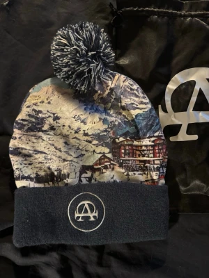 Mössa med vintermotiv och tofs - Säljer en snygg mössa med vinterlandskap och fjällmotiv i blå, vit och beige ton. Mössan har en stor svartvit tofs på toppen och vikt kant med ett broderat A-logotyp framtill. Perfekt för dig som vill sticka ut i backen eller på stan.