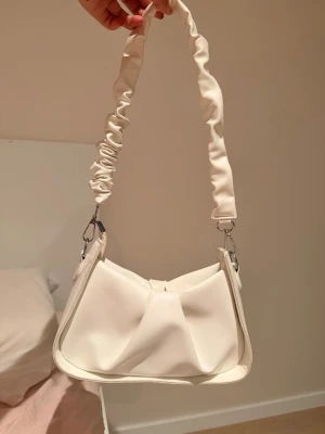 Vit axelremsväska med scrunchie-band - Trendig vit handväska i mjukt konstläder med unik scrunchie-inspirerad axelrem. Väskan har en modern, veckad design och silverfärgade metalldetaljer. Perfekt accessoar för att lyfta din outfit med en stilren och ungdomlig touch.