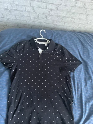 Svart piké från Calvin Klein M - Snygg svart pikétröja från Calvin Klein i storlek M. Skjortan har små vita loggor över hela tyget, klassisk krage och knappar vid halsen. Perfekt för dig som gillar stilrena och moderna plagg med diskreta detaljer.