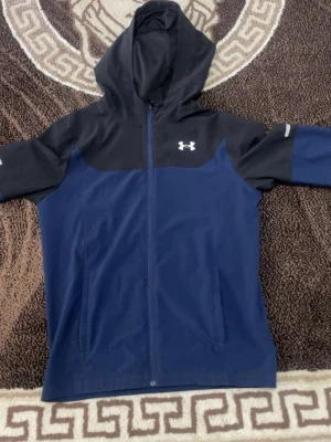 Blå och svart jacka från Under Armour - Snygg jacka från Under Armour i blått och svart med dragkedja framtill och två sidofickor. Hoodien har en stor huva och logga på bröstet. Perfekt för dig som gillar sportig stil och vill ha något bekvämt och funktionellt.