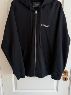Svart zip hoodie från Replay - Snygg svart hoodie från Replay i storlek XL. Dragkedja framtill, två fickor och broderad Replay-logga på bröstet. Klassisk huva med dragsko och mjukt material som känns skönt mot huden. Perfekt för en avslappnad streetwear-look.