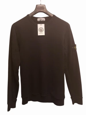 Svart sweatshirt från Stone Island - Stilren svart sweatshirt från Stone Island med klassisk rund halsringning och ikonisk logopatch på vänster ärm. Tröjan har ribbade muddar vid ärmslut och nederkant, perfekt för en clean och avslappnad look.