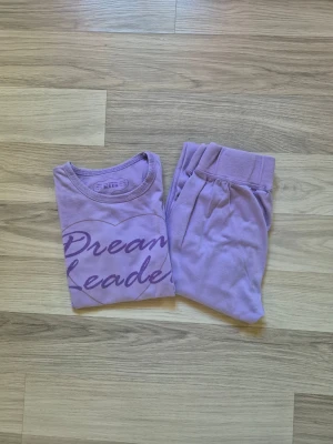 Lila pyjamas med text Dream Leader - Mysig lila pyjamas med långärmad tröja och matchande byxor. Tröjan har trycket 'Dream Leader' i mörkare lila och ett hjärta på bröstet. Tillverkad i mjukt material, perfekt för chillkvällar eller sovmorgnar.