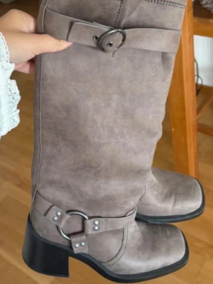 Grå höga boots med spänne och klack - Snygga höga boots i grått med markerad klack och coola spänndetaljer i silver. Stövlarna har rund tå, dekorativa remmar och ringar runt ankeln samt upptill. Tillverkade i ett mockaliknande material som ger en trendig look.