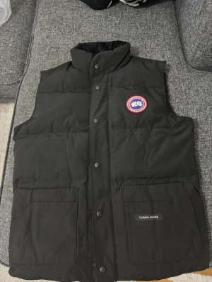 Svart dunväst från Canada Goose - Snygg svart dunväst från Canada Goose med hög krage och tryckknappar framtill. Västen har två stora fickor framtill, klassisk logotyp på bröstet och en stilren design. Perfekt för lager-på-lager och håller dig varm under kyliga dagar.
