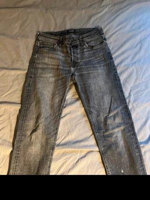 Grå tvättade raka jeans - Säljer ett par grå tvättade jeans med rak passform och klassisk femficksdesign. Jeansen har subtila slitningar och kontrastsömmar, samt en svart patch bak i midjan. Perfekta för en avslappnad och trendig look.