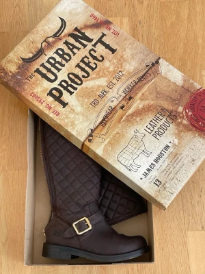Bruna quiltade ridstövlar med spänne - Snygga bruna ridstövlar från The Urban Project i quiltat läder med dekorativt spänne och dragkedja på sidan. Stövlarna har en robust sula och klassisk form som passar perfekt till höstens och vinterns outfits.