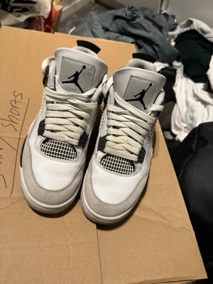Vita Air Jordan 4 Retro sneakers - Säljer ett par klassiska Air Jordan 4 Retro sneakers i vitt med svarta och grå detaljer. Skorna har meshpaneler, synlig Air-sula och ikonisk Jumpman-logga på plösen. Perfekta för dig som vill ha en stilren och sportig look.