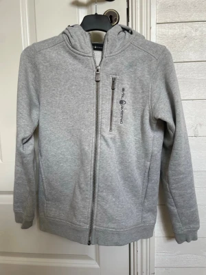 Ljusgrå hoodie från Sail Racing - Ljusgrå hoodie med dragkedja från Sail Racing. Tröjan har huva, bröstficka med dragkedja och logga på vänster sida. Tillverkad i mjukt sweatshirtmaterial och har ribbade muddar vid ärmar och nederkant. Perfekt för en avslappnad och sportig stil. Det är storlek 150 men gick inte o välja det men det är så ni vet
