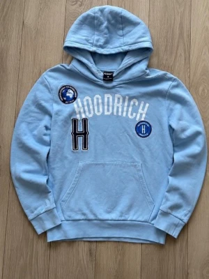 Ljusblå hoodie från Hoodrich - Ljusblå hoodie med huva från Hoodrich. Framsidan har vita och svarta tryck och broderade patches, inklusive texten 'HOODRICH' och två runda märken. Klassisk känguruficka och ribbade muddar. Perfekt för en avslappnad streetwear-look. Storlek M