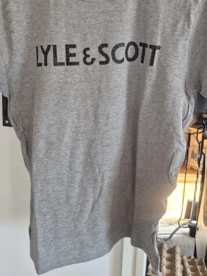 Grå t-shirt från Lyle & Scott - Säljer en enkel och stilren grå t-shirt från Lyle & Scott med svart logga tryckt på bröstet. T-shirten har klassisk passform och rund hals. Perfekt till jeans eller shorts för en clean look.