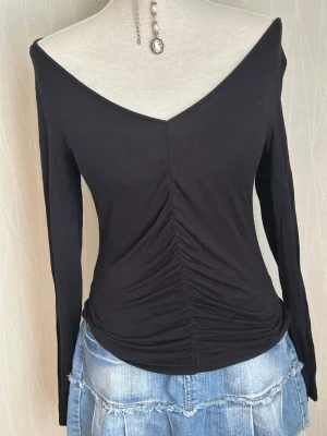 Svart långärmad topp med v-ringning - Trendig svart topp med lång ärm och djup v-ringning. Toppen har rynkade detaljer framtill som ger en snygg siluett. Tillverkad i mjukt material, perfekt att styla med jeans eller kjol för en cool look.