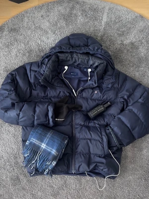 Mörkblå pufferjacka från Gant - Snygg mörkblå pufferjacka från Gant med huva och dragkedja framtill. Jackan har en diskret logga på bröstet och quiltad design som ger en modern look. Perfekt för kalla vinter och höstdagar. Storlek 176 / XS men passar någon runt 170cm. Nypris 1499. Endast jacka till salu.