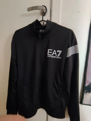 EA7 hoodie  - Snygg svart ziptröja från EA7 Emporio Armani med vit logga på bröstet och vita ränder på ena ärmen. Tröjan har hög krage, dragkedja framtill och två fickor med dragkedja. Perfekt för en sportig och stilren look.