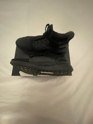 Adidas Ultra Boost 04 - Säljer ett par helsvarta sneakers med stickad ovandel och grov sula. Skorna har en sportig look med plastdetaljer på sidan och snörning. Perfekta för dig som gillar en clean och modern stil.
