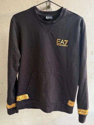 Svart sweatshirt från EA7 Emporio Armani - Svart sweatshirt med gult EA7 Emporio Armani-tryck på bröstet och detaljer på muddar och nederkant. Klassisk rund halsringning och magficka framtill. Tillverkad i mjuk bomullsblandning för en skön och avslappnad look.