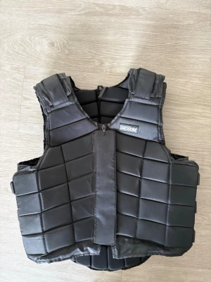 Svart ridväst från Jacson - Säljer en svart ridväst från Jacson med vadderad design och dragkedja framtill. Västen har justerbara axelband och sidoremmar för bästa passform. Perfekt för ridning 