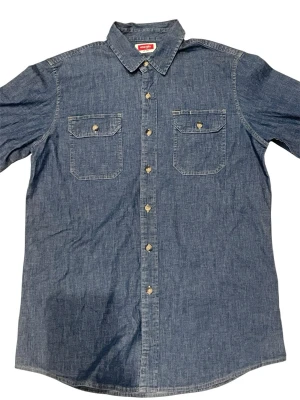 Blå jeansskjorta från Wrangler - Klassisk blå jeansskjorta från Wrangler med två bröstfickor och snygga knappar framtill. Skjortan har lång ärm och en rak passform. Perfekt för dig som gillar en tidlös och avslappnad stil.