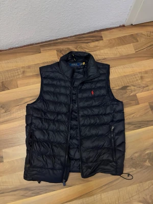 Svart dunväst från  Ralph Lauren - Snygg svart dunväst från Polo Ralph Lauren med hög krage och dragkedja framtill. Västen har två fickor med dragkedja och den klassiska röda loggan broderad på bröstet. Perfekt för lager på lager och streetwear-stil.