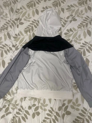 Nike vindjacka med huva grå/vit - Nike vindjacka i grått och vitt med svart panel över axlarna. Jackan har huva, långa ärmar och resår i nederkant. Tillverkad i lätt och vindtätt material, perfekt för blåsiga dagar. Snygg och sportig design som passar till streetwear.