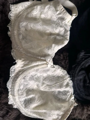 Vit spets-bh med rosett - Säljer en vit bh med vacker spets och liten rosett framtill. Bh:n har bygel och breda axelband för extra stöd. Materialet består av 86% nylon och 14% elastan vilket gör den både mjuk och stretchig. Perfekt för dig som gillar romantiska detaljer.