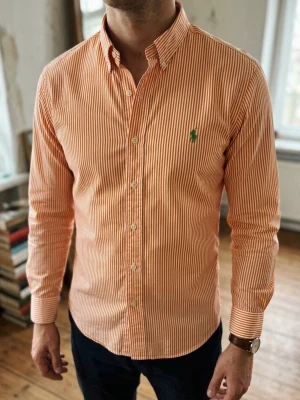 Randig orange skjorta från Polo Ralph Lauren (Slim Fit) - Säljer en snygg orange och vit randig skjorta från Ralph Lauren. Skjortan har klassisk krage, långa ärmar och knappar framtill. Den ikoniska gröna loggan är broderad på bröstet. Perfekt för dig som vill ha en stilren och fräsch look. Storlek M. Snabb Leverans!