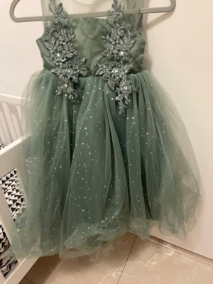 Grön glittrig prinsessklänning - Snygg grön kortklänning med glittrande tyllkjol och detaljerad spetsbrodyr med pärlor och blommor på överdelen. Klänningen har ärmlös design och ger en riktigt sagolik känsla med sitt skimrande material och vackra dekorationer.