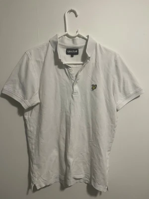 Vit pikétröja från Lyle & Scott - Klassisk vit pikétröja från Lyle & Scott med korta ärmar och krage. Tröjan har knappar vid halsen och den ikoniska gula örnloggan broderad på bröstet. Perfekt för en stilren och avslappnad look.