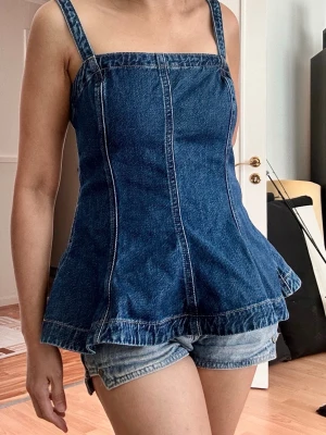 Jeanslinnetopp från H&M Divided - Säljer en snygg jeanslinnetopp från H&M Divided i mörkblå denim. Toppen har breda axelband, markerade sömmar och en lätt utsvängd nederkant som ger en cool siluett. Perfekt till sommaren och enkel att styla med shorts eller kjol.