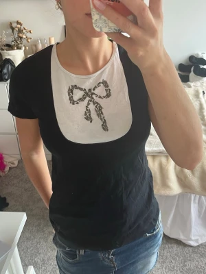 Svart t-shirt med rosettdetalj - Snygg svart t-shirt med vit detalj och en rosett av ”metall” skärvor. T-shirten har rund halsringning och korta ärmar. Perfekt för dig om du vill ha en unik twist på din vardagsstil.